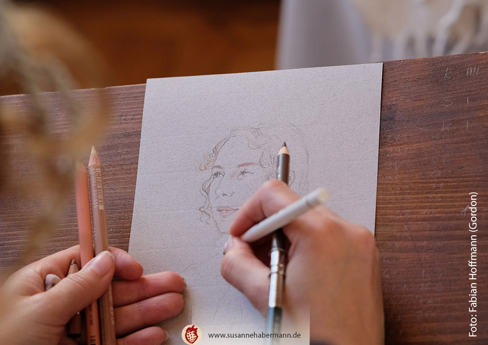Porträtzeichnen - Künstlerin beim Zeichnen auf einem Zeichenbrett, im Vordergrund eine Portraitzeichnung im Entstehen - Porträtzeichnen auf Veranstaltungen mit Susanne Habermann - Foto: Fabian Hofmann